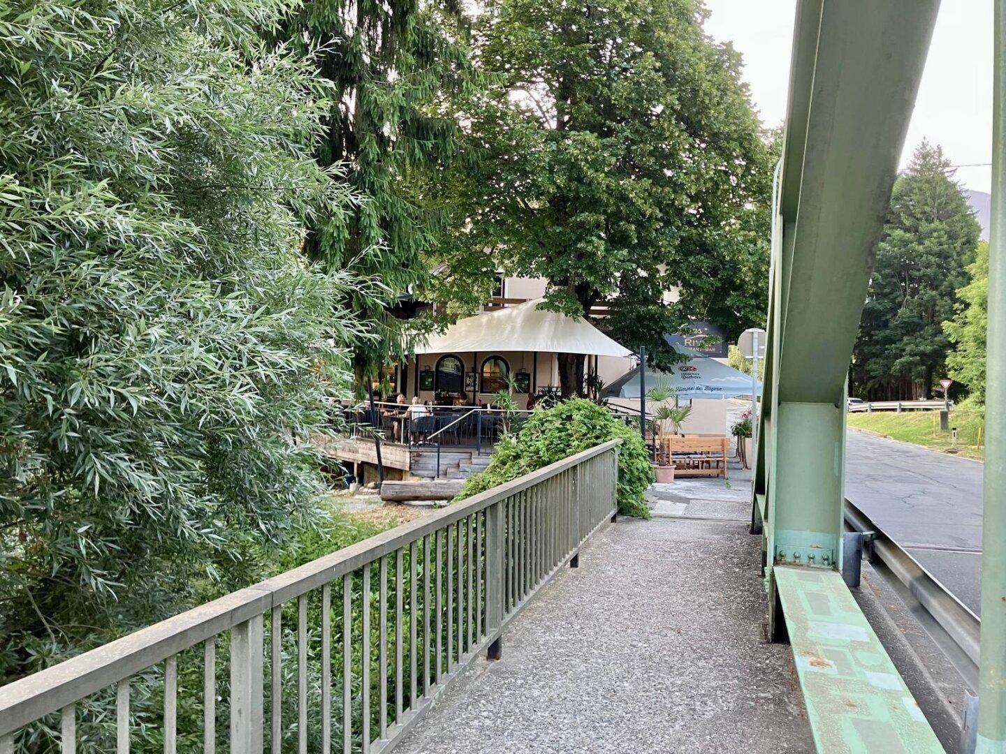 Blick vom Gehweg auf das Restaurantgebäude, umgeben von üppiger Natur und einem Fluss.