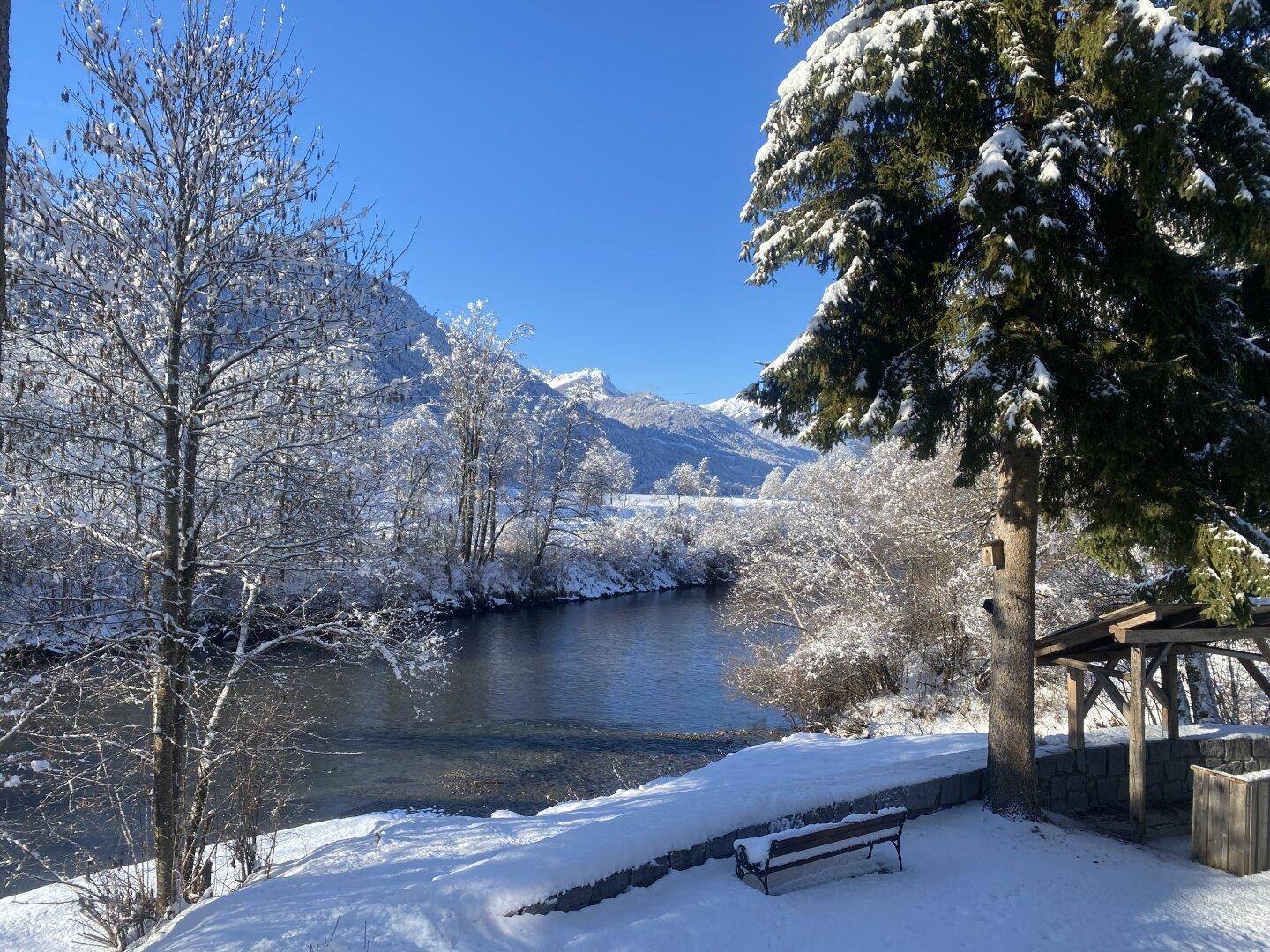 Idyllische Winterlandschaft mit schneebedeckten Bäumen, einem Fluss und Bergen im Hintergrund.