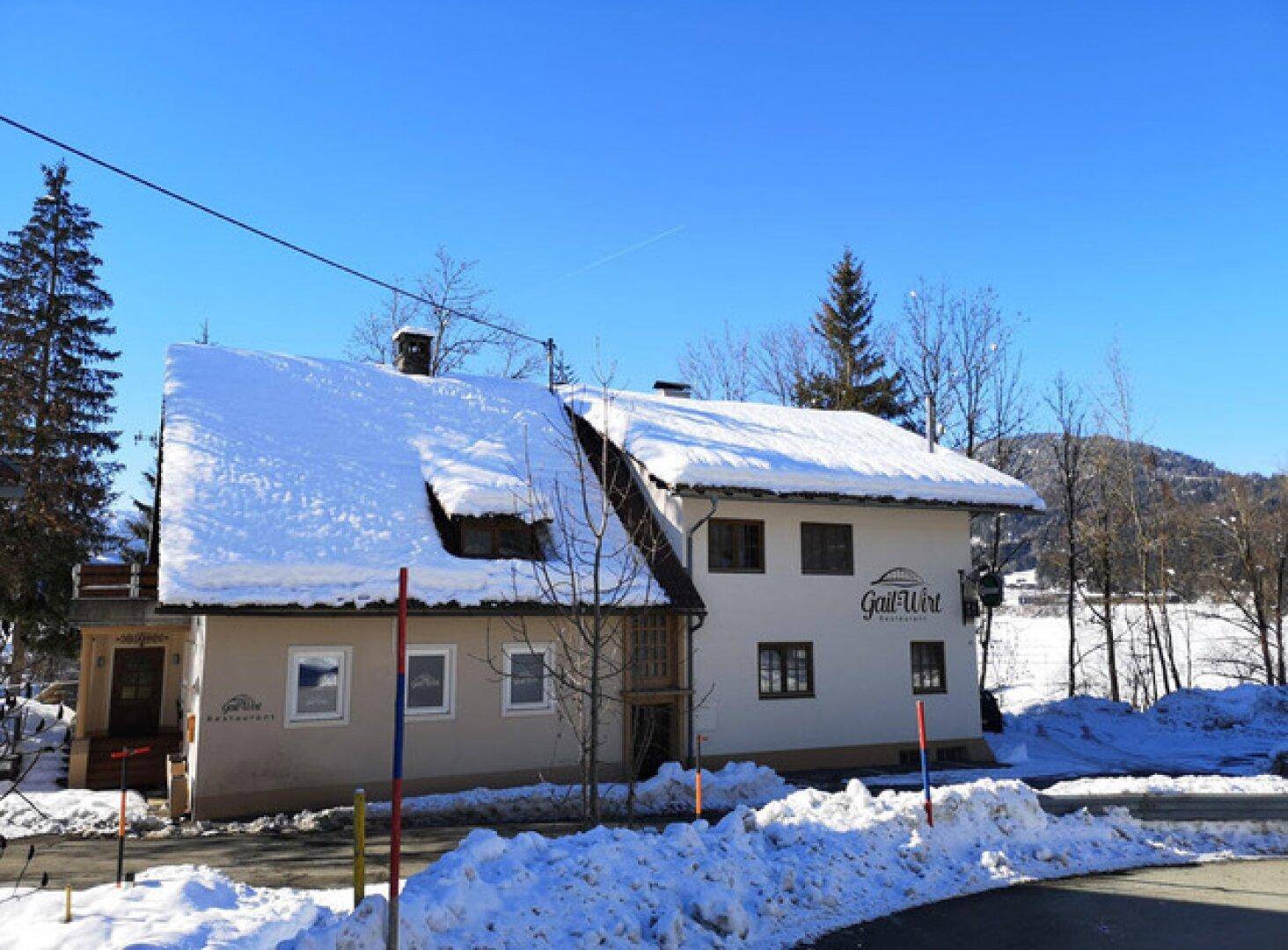 Traditionelles Gebäude mit schneebedecktem Dach in winterlicher Landschaft, umgeben von Bäumen.