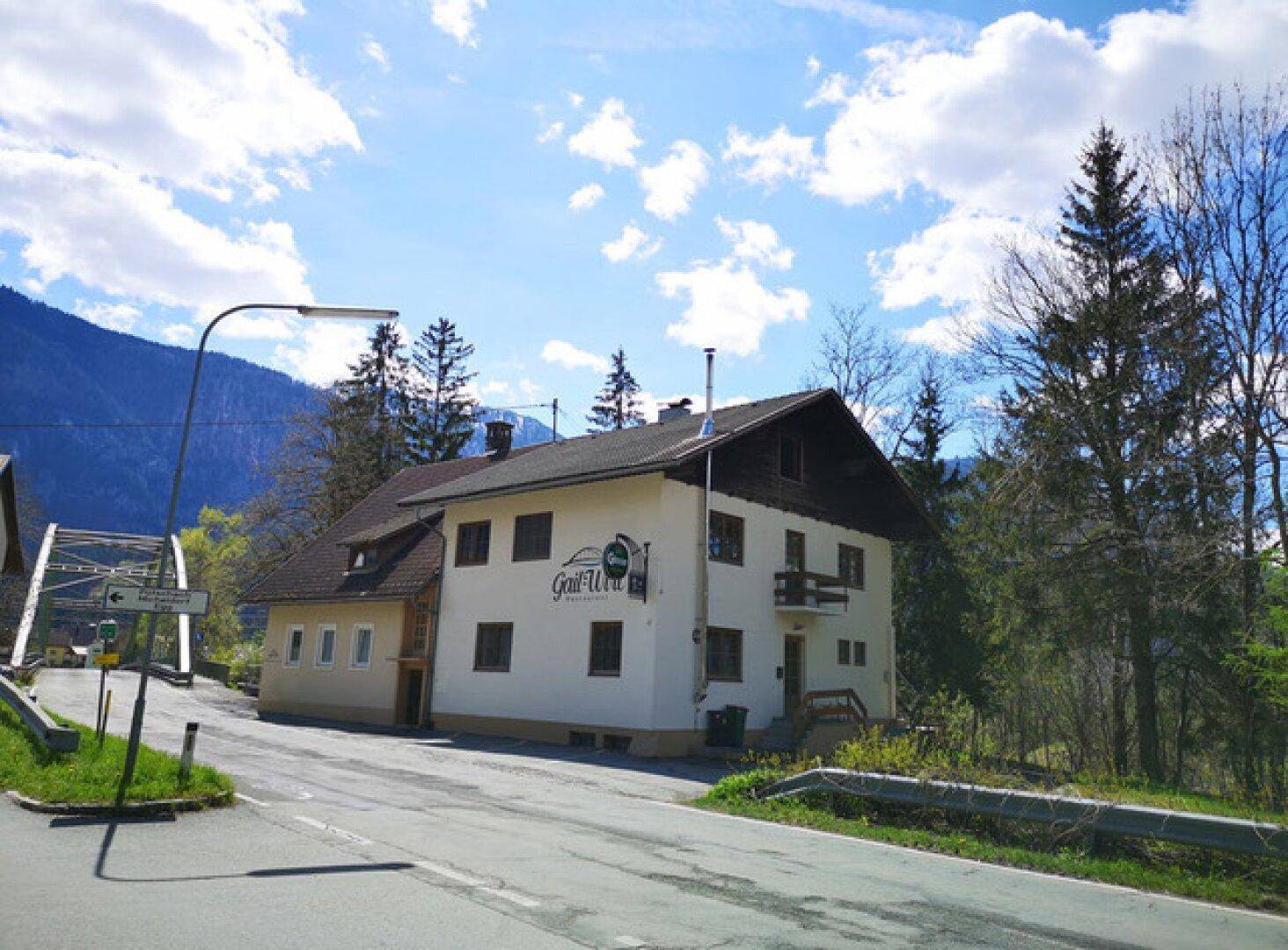 Traditionelles Gebäude mit Satteldach in grüner Umgebung, umgeben von Bäumen und Bergen.