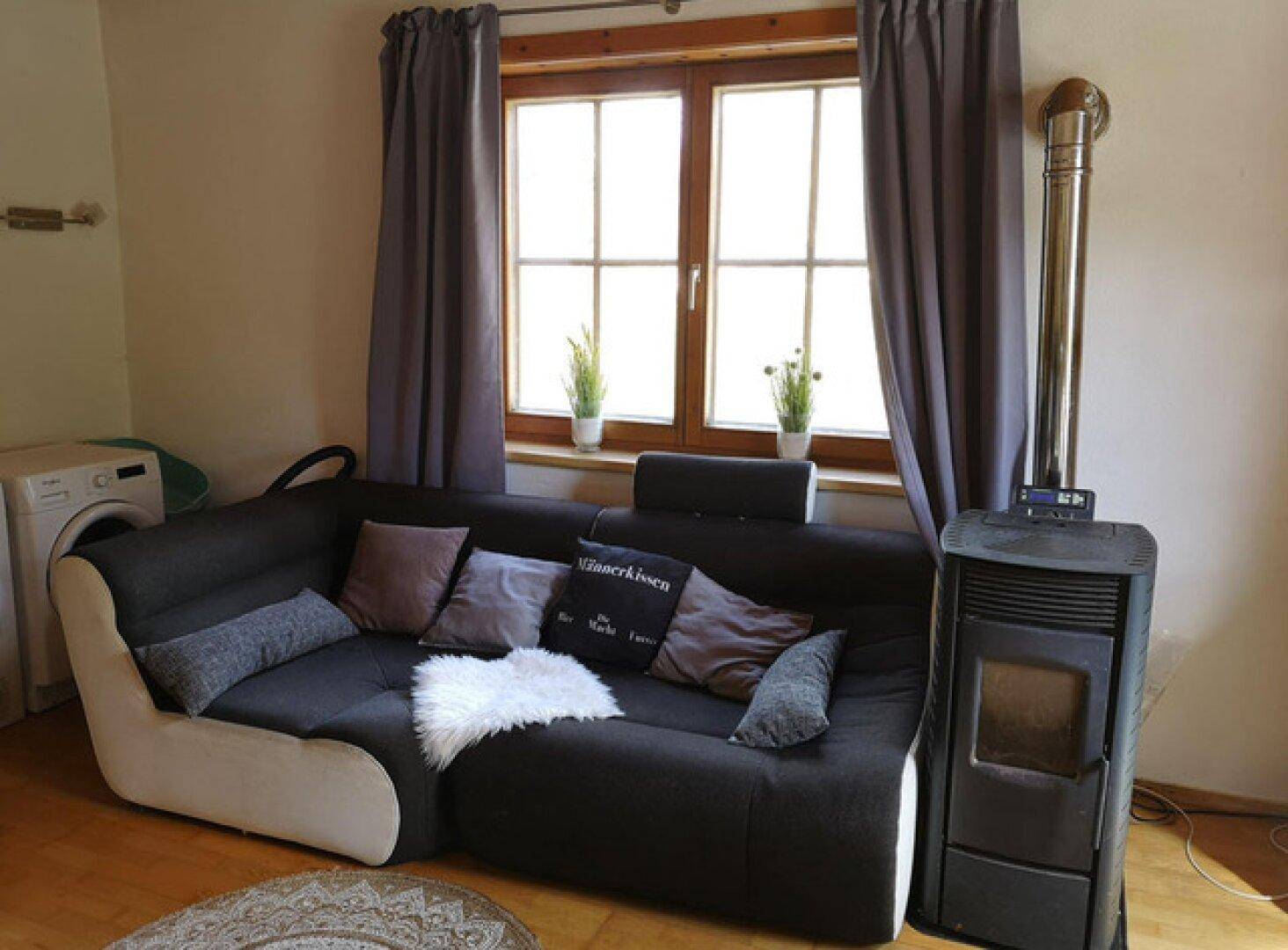 Detailansicht des Wohnzimmers mit bequemer Eckcouch und einem modernen Holzofen am Fenster.