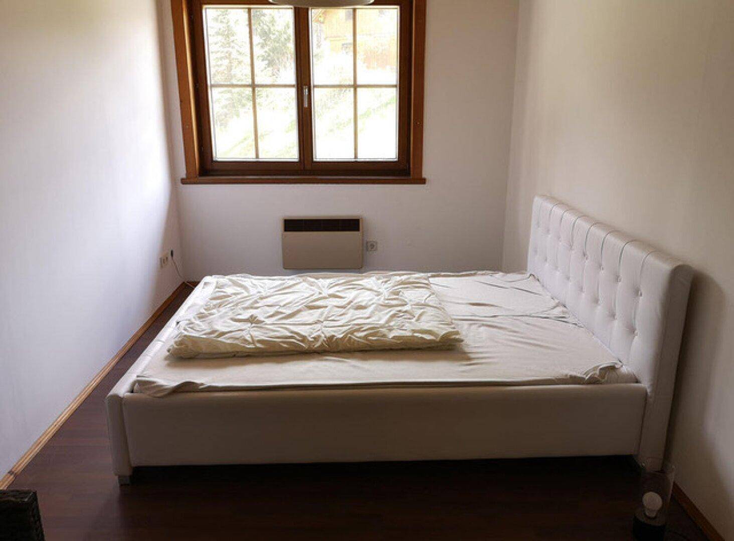 Helles Schlafzimmer mit weißem Doppelbett, Holzboden und einem Fenster mit Blick ins Grüne.