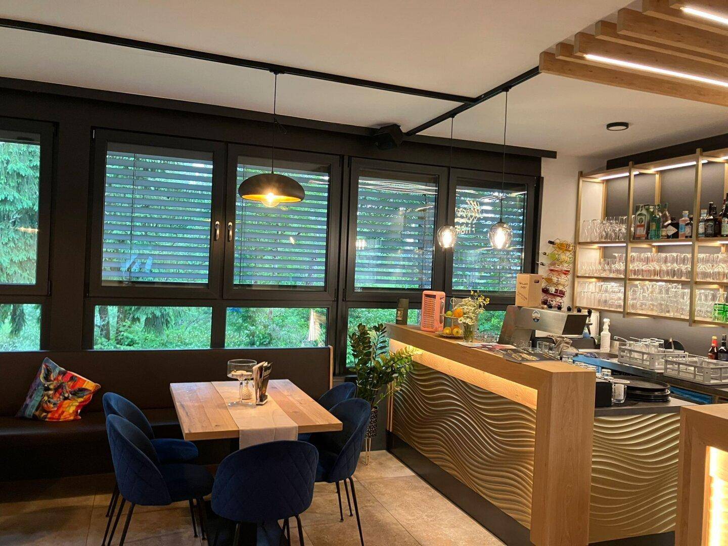 Stilvolle Bar mit Holztheke und beleuchteten Regalen neben einem Esstisch am Fenster mit Blick ins Grüne.