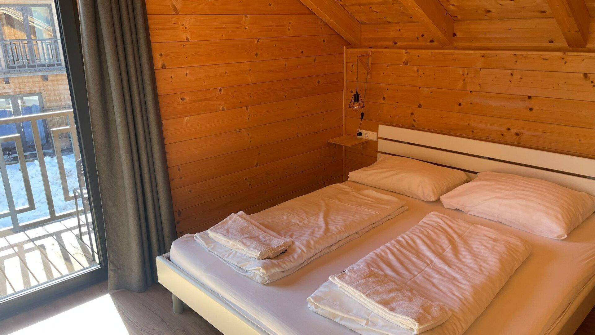 Helles Schlafzimmer mit Holzwänden, Doppelbett und direktem Zugang zum Balkon, unter einem Dachschrägen.