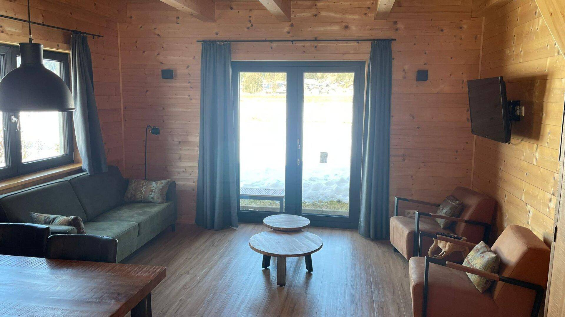 Heller Wohnbereich mit Holzwänden, bequemer Couch und großen Fenstertüren, die viel Tageslicht hereinlassen.