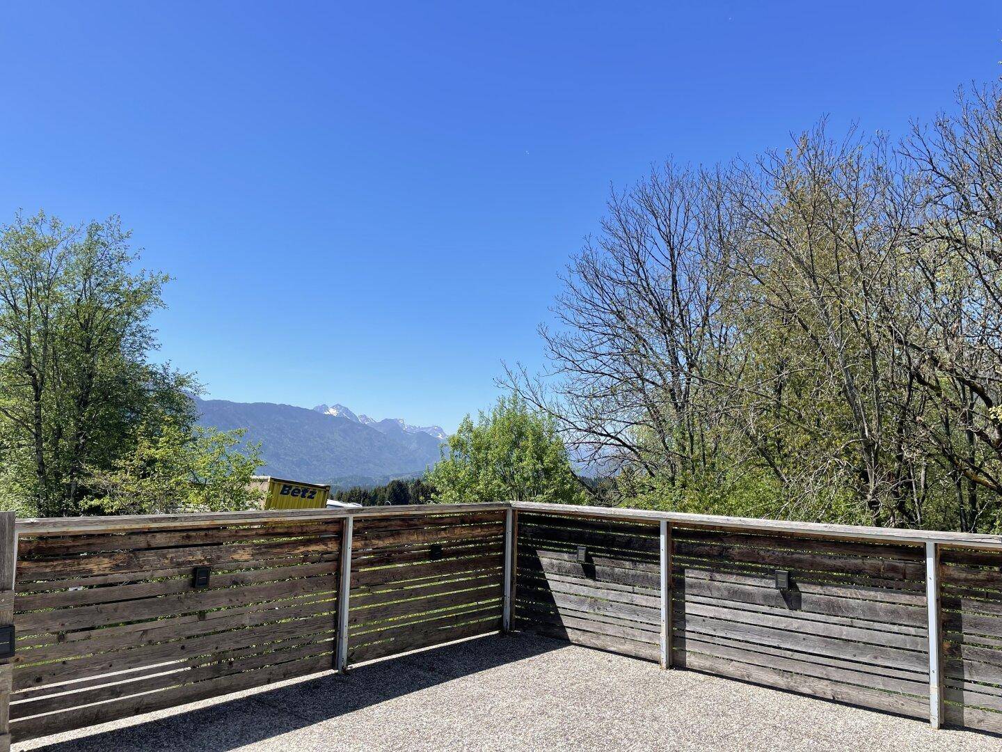 Geräumige Terrasse mit Holzzaun und weitem Blick auf die umliegende Berglandschaft und grüne Bäume.