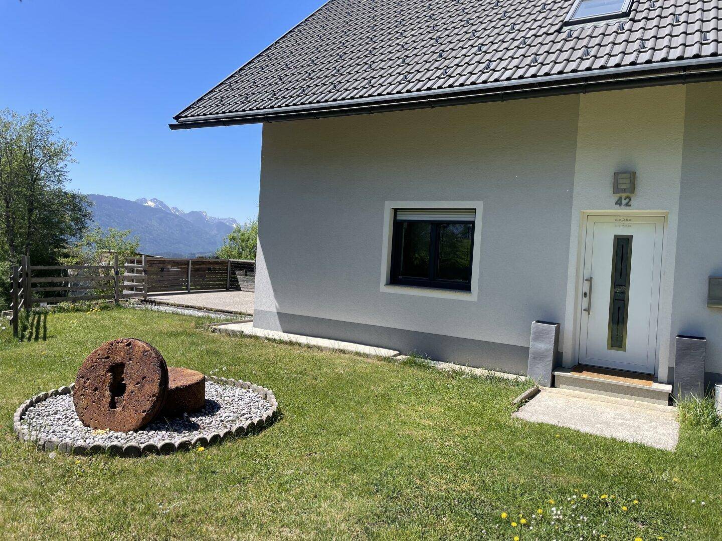 Eingangsbereich des Hauses mit weißer Tür, umgeben von Rasenfläche und einem dekorativen Steinelement, mit Bergblick.