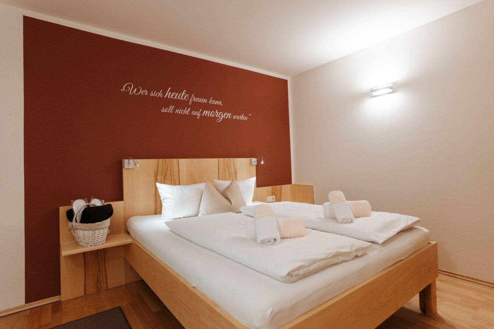 Gemütliches Schlafzimmer mit Doppelbett, Holzelementen und einer roten Akzentwand mit Schriftzug.