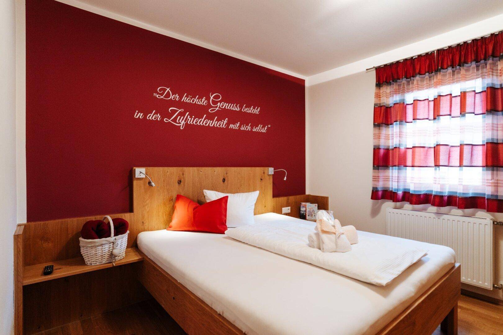 Schlafzimmer mit Doppelbett, roter Akzentwand und Fenster mit gestreiften Vorhängen.