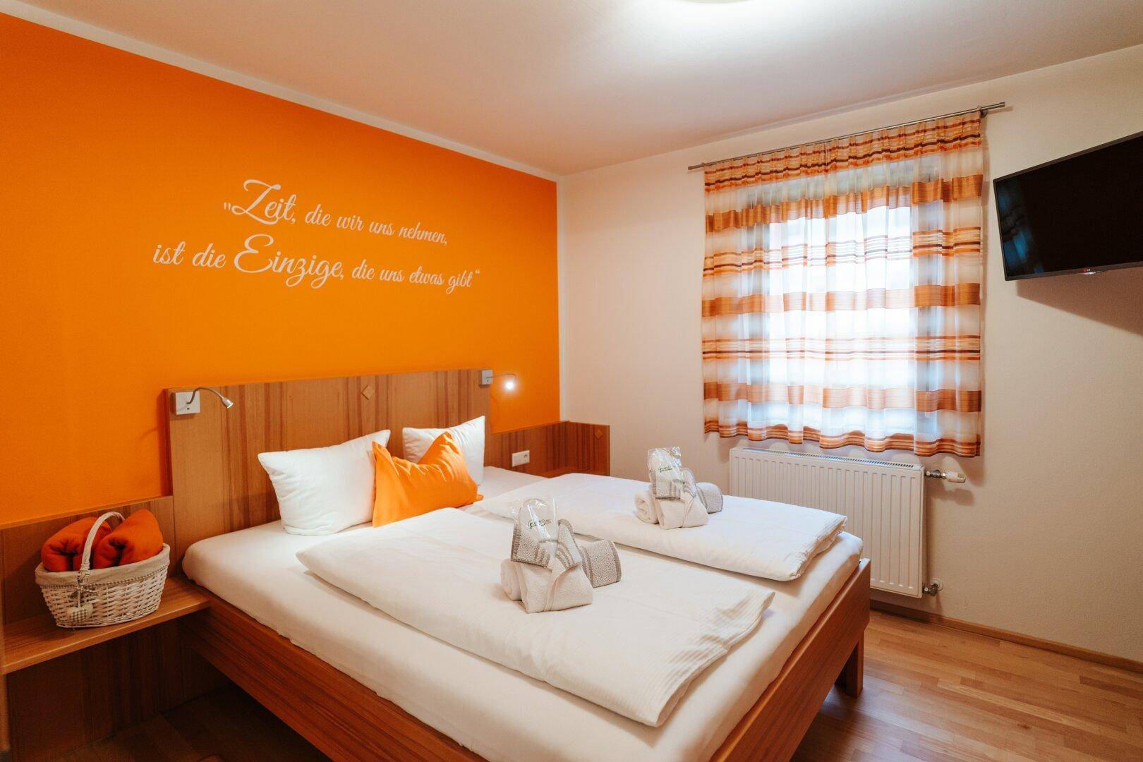 Schlafzimmer mit Doppelbett, orangefarbener Akzentwand und Holzelementen.