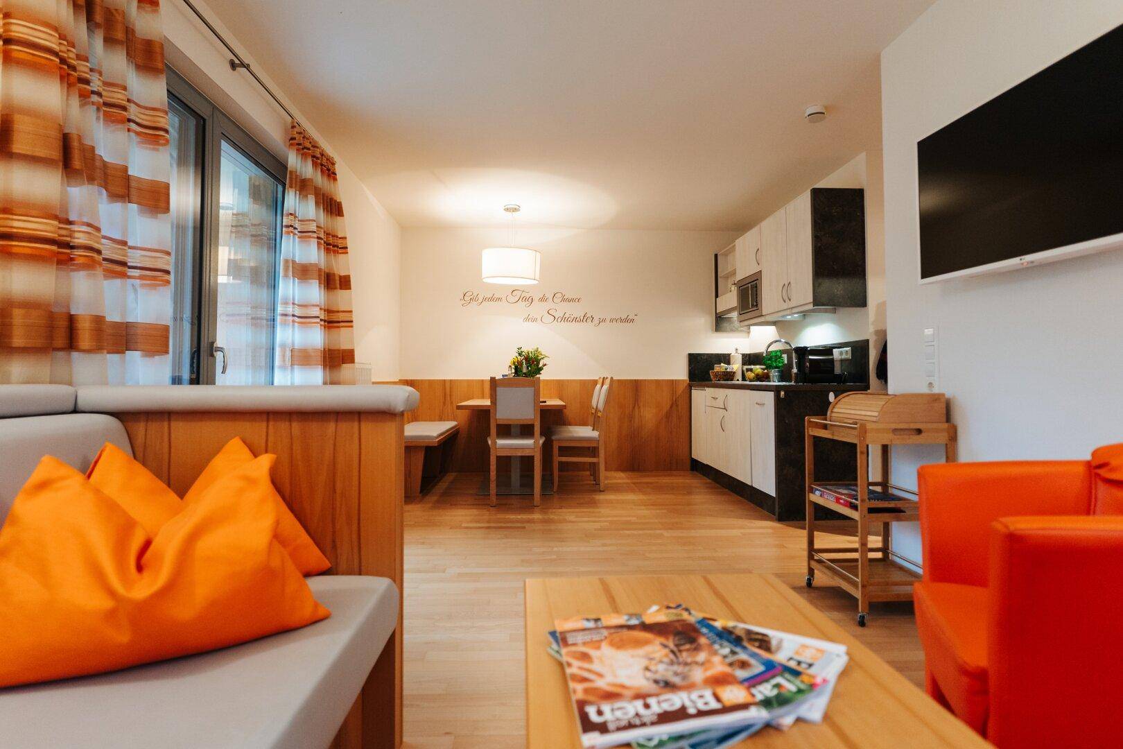 Heller Wohn- und Essbereich mit Küchenzeile, Holzboden und orangefarbenen Akzenten.
