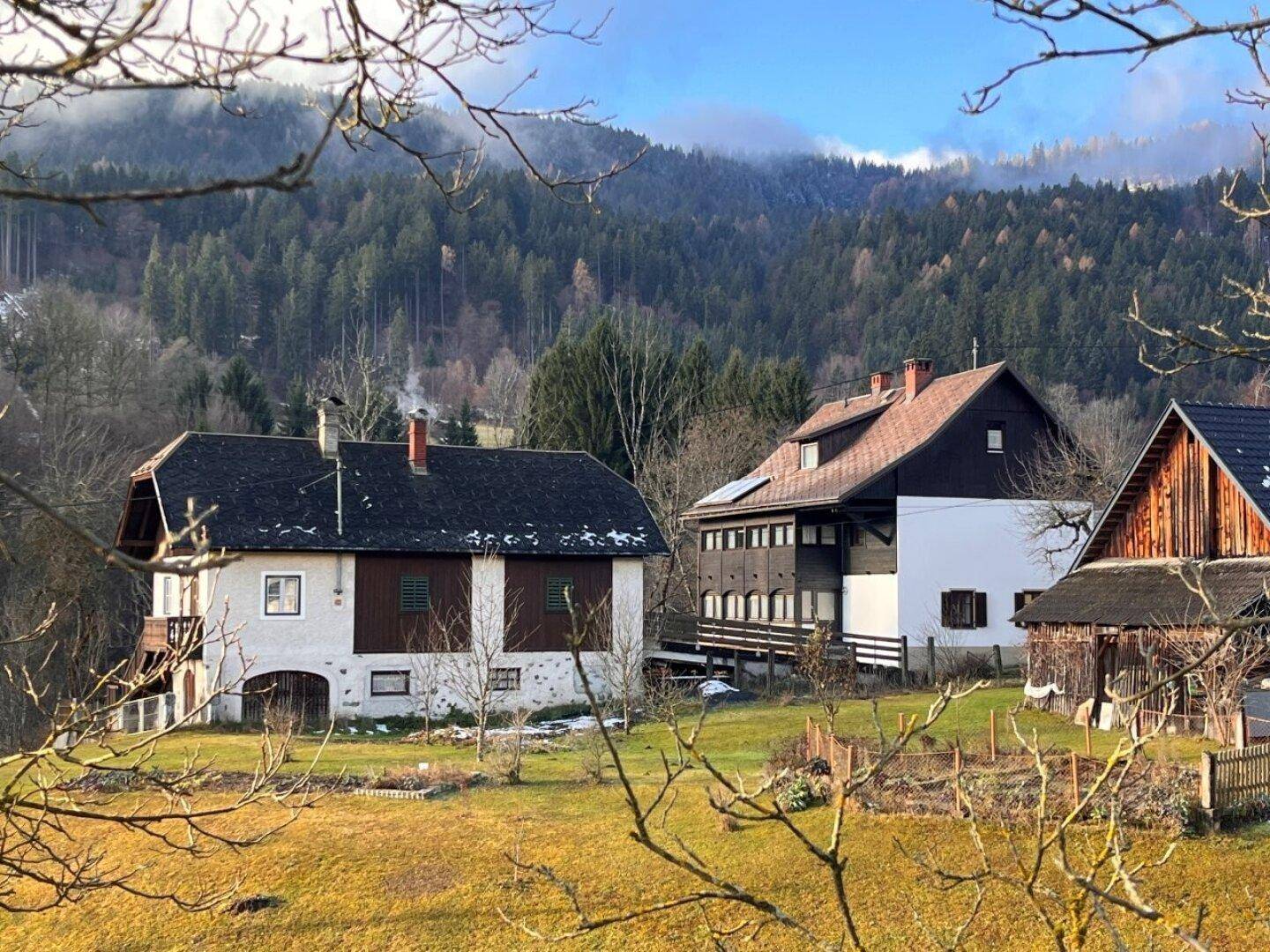 Traditionelle Häuser in einer ländlichen Umgebung mit bewaldeten Bergen im Hintergrund unter blauem Himmel.
