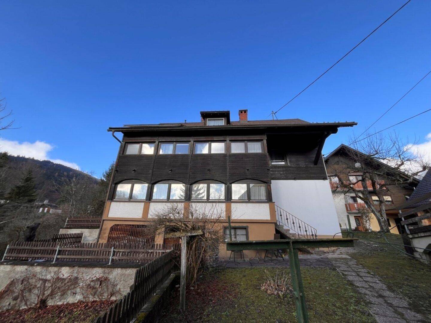 Traditionelles Haus mit dunkler Holzfassade und vielen Fenstern, eingebettet in eine Hanglage.
