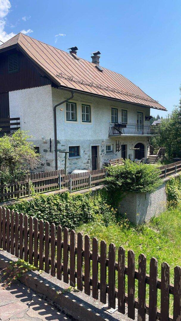 Traditionelles Haus mit Balkon, Holzzaun und üppiger Vegetation unter blauem Himmel.