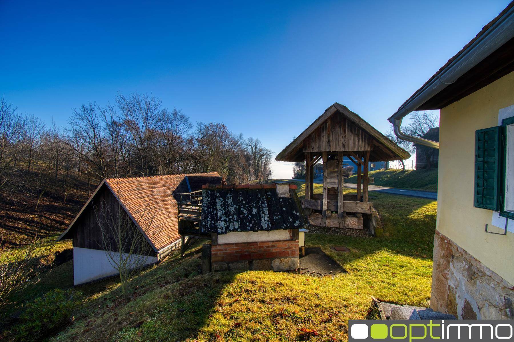 Historischer Holzbrunnen und Nebengebäude in ländlicher Umgebung unter strahlend blauem Himmel.