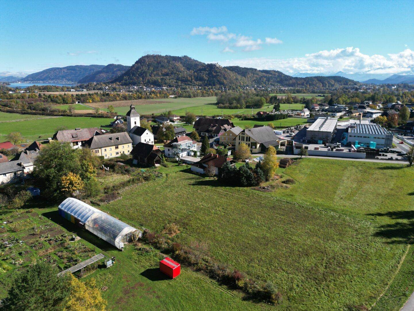 Weitwinkelaufnahme eines grünen Feldes mit Dorfbebauung und einem See im Hintergrund, umgeben von Hügeln.