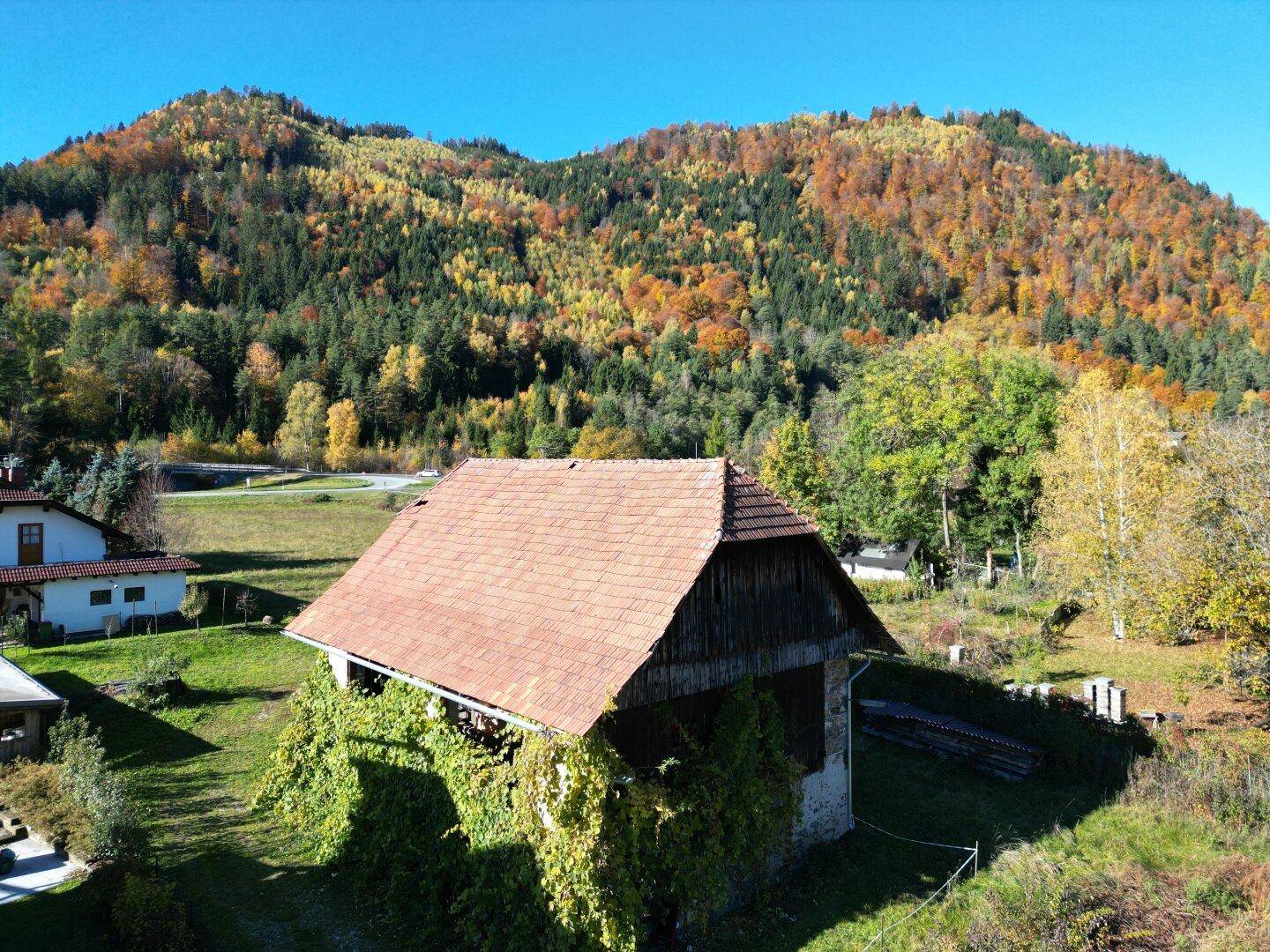 Luftaufnahme eines alten Scheunengebäudes mit Ziegeldach, umgeben von üppigem Grün und einem herbstlichen Wald.