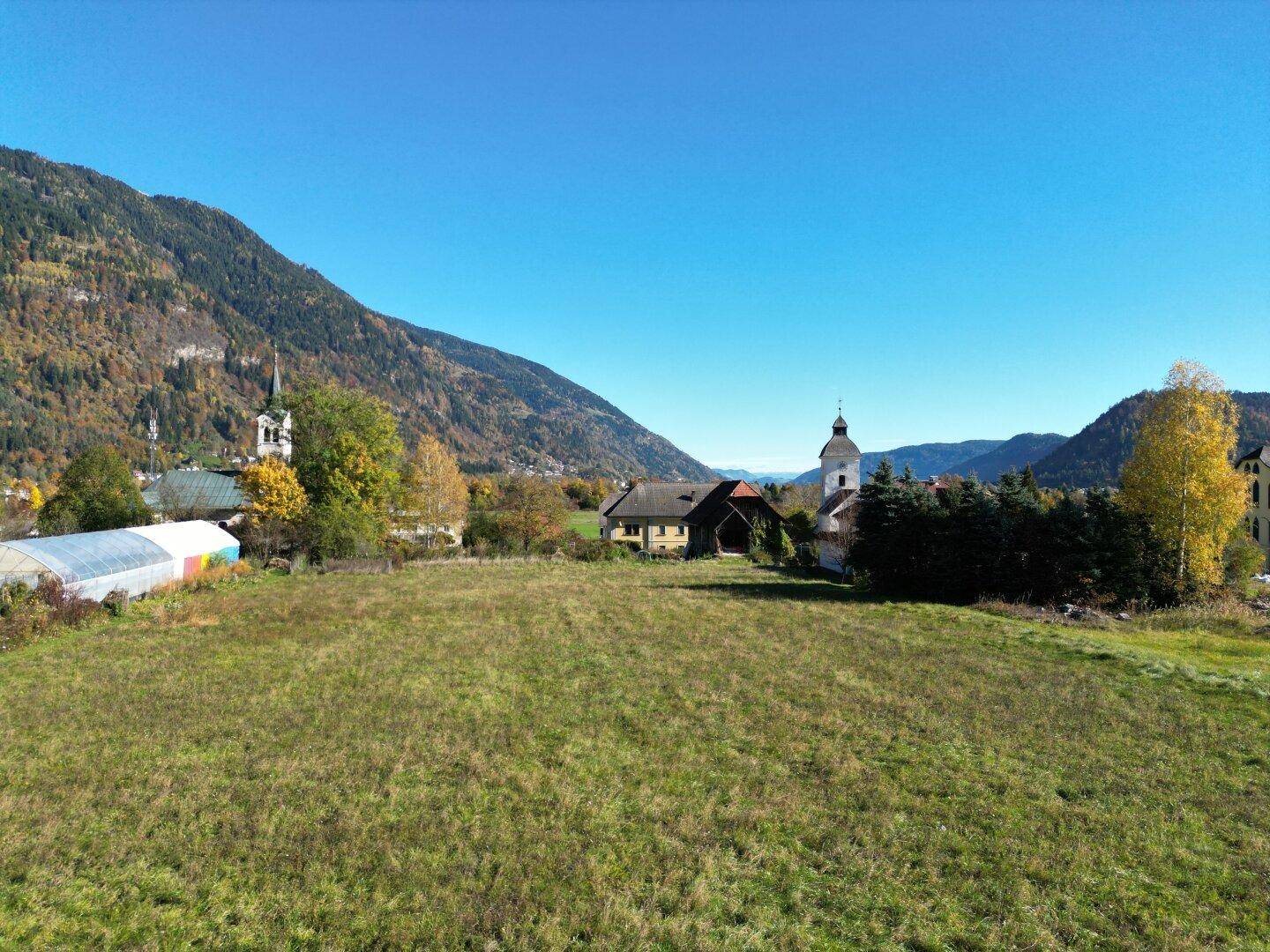 Panoramablick auf ein großes grünes Grundstück, umgeben von ländlicher Bebauung und einer beeindruckenden Bergkulisse.