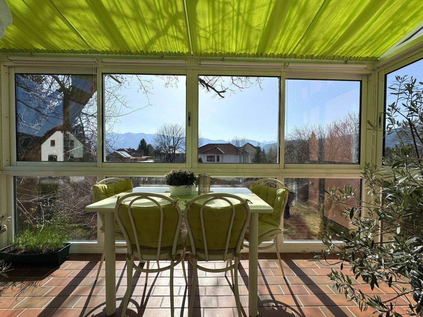 Lichtdurchfluteter Wintergarten mit Terracotta-Fliesenboden, Gartenmöbeln und Bergblick.