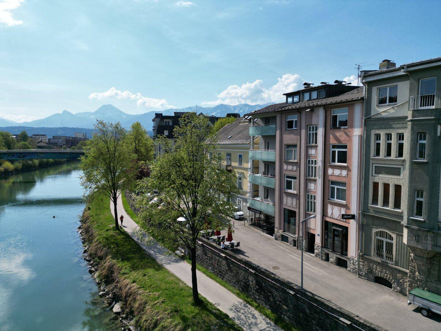 Flussufer mit Gebäuden, Bäumen und einer Brücke im Hintergrund, umgeben von Bergen.