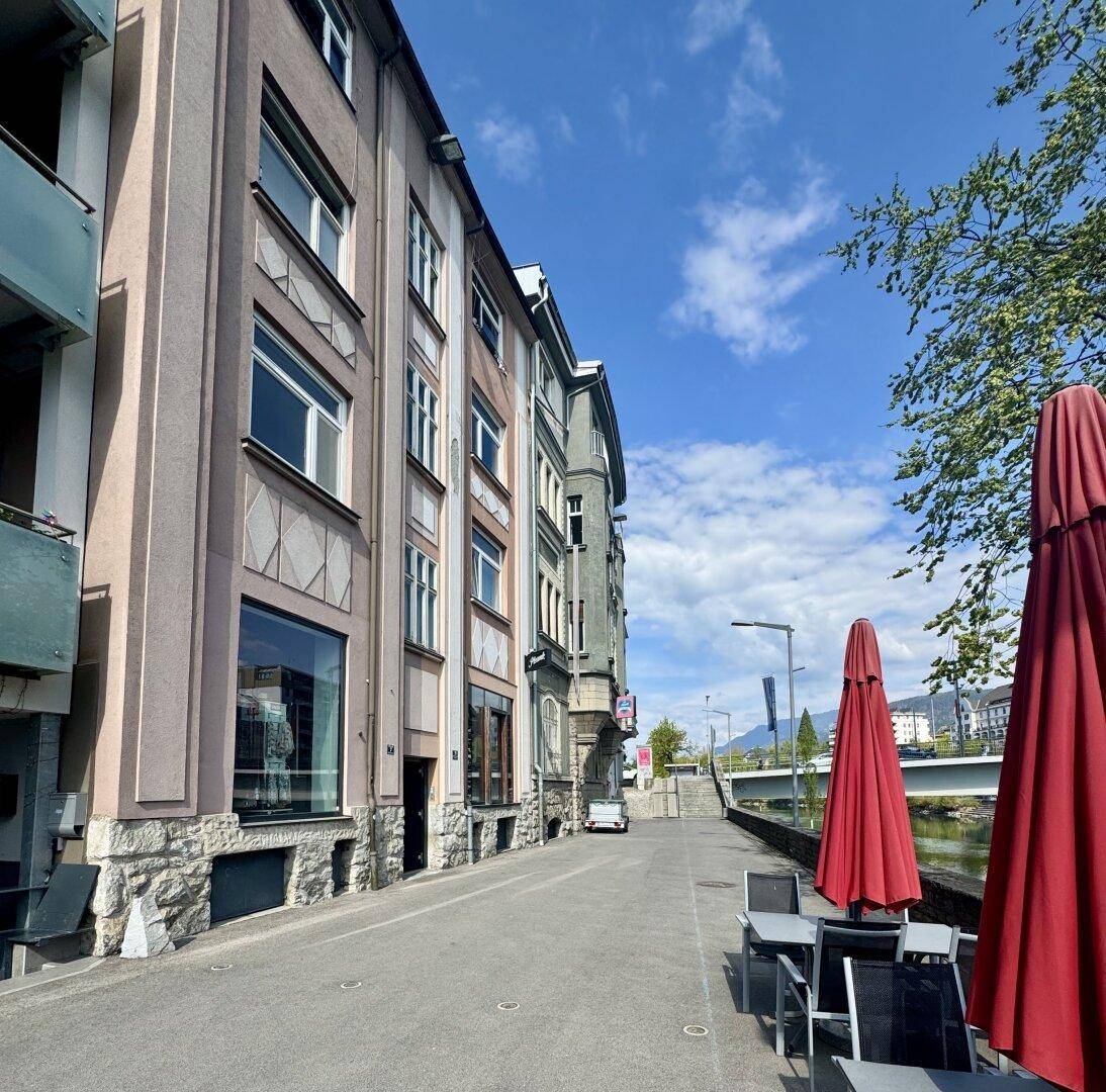 Straßenszene mit Gebäudefassaden, Außengastronomie und einem Fluss im Hintergrund unter blauem Himmel.