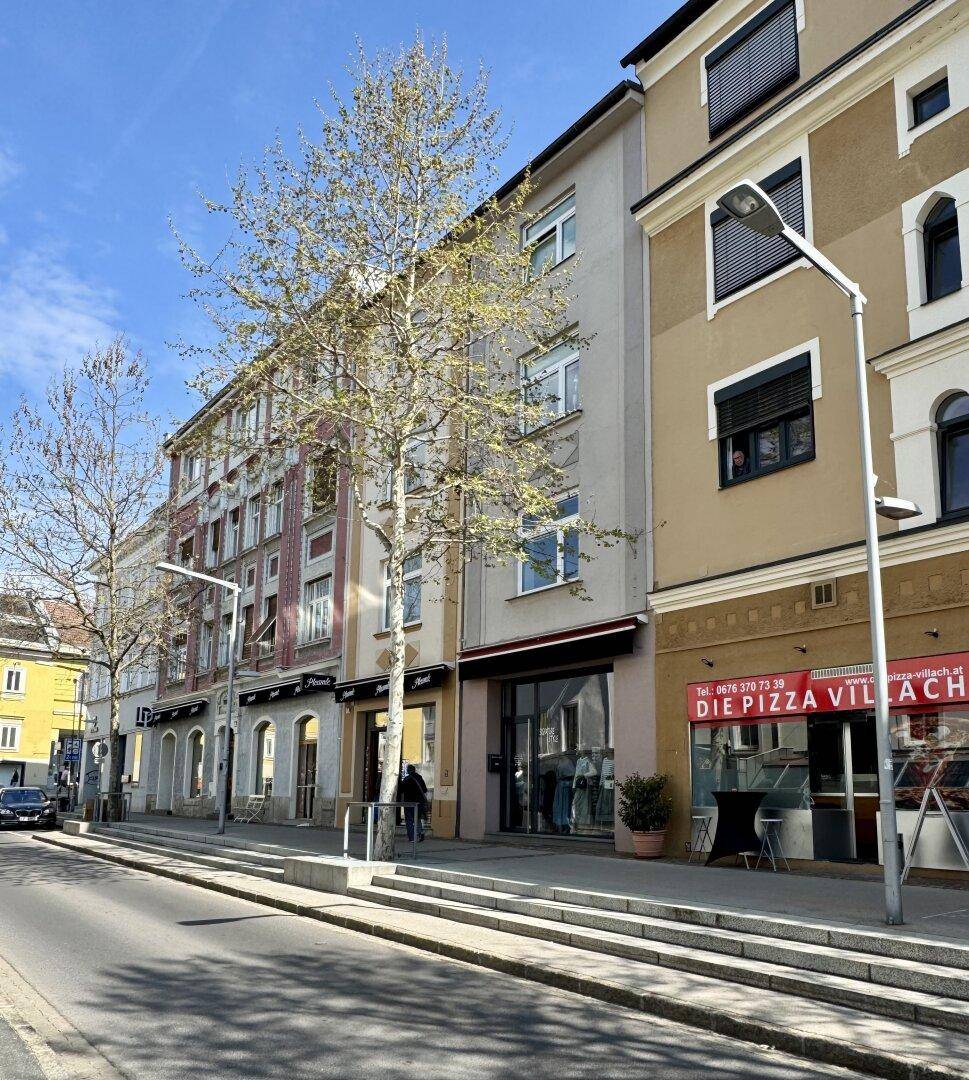Eine Reihe von Gebäuden entlang einer Straße mit Geschäften im Erdgeschoss und Bäumen.