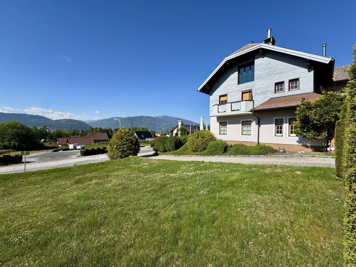 Seitenansicht des Gebäudes mit Balkon und großer Rasenfläche, umgeben von grüner Landschaft und Bergen.