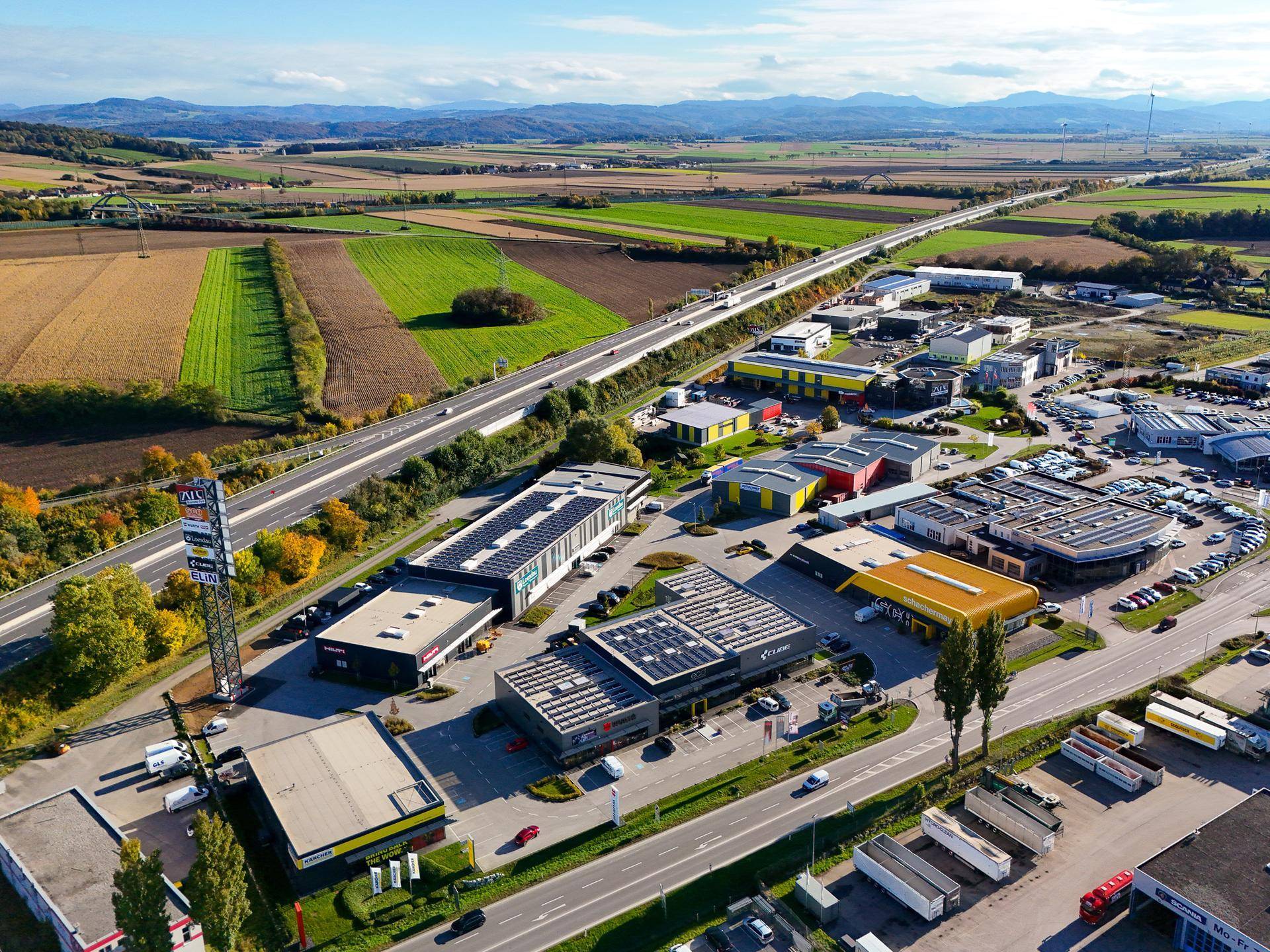 Luftaufnahme eines Gewerbeparks mit verschiedenen Gebäuden, Straßen und umliegender Landschaft mit Bergen im Hintergrund.