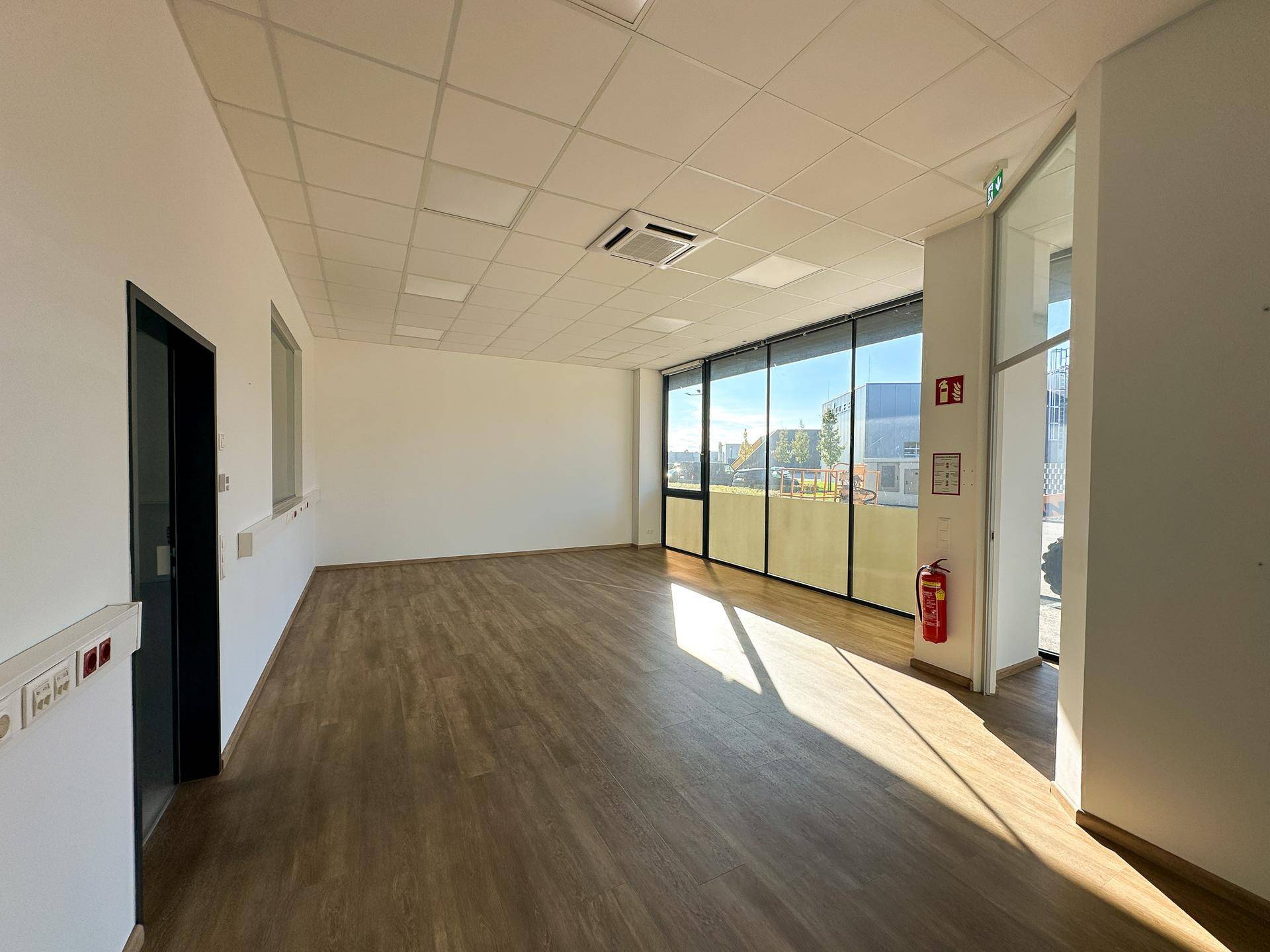 Heller, leerer Gewerberaum mit großen Fenstern, Holzboden und abgehängter Decke mit integrierter Beleuchtung.