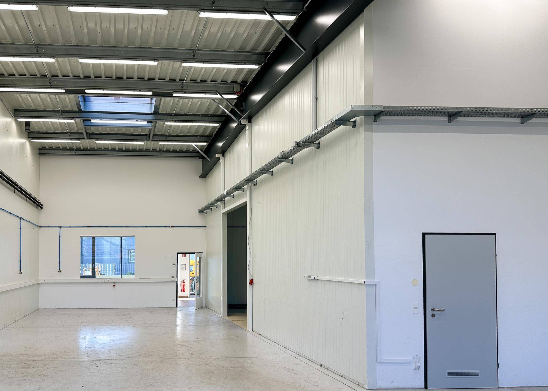Weitläufige Lagerhalle mit hoher Decke, sichtbaren Trägern und industrieller Beleuchtung sowie einem Fenster.