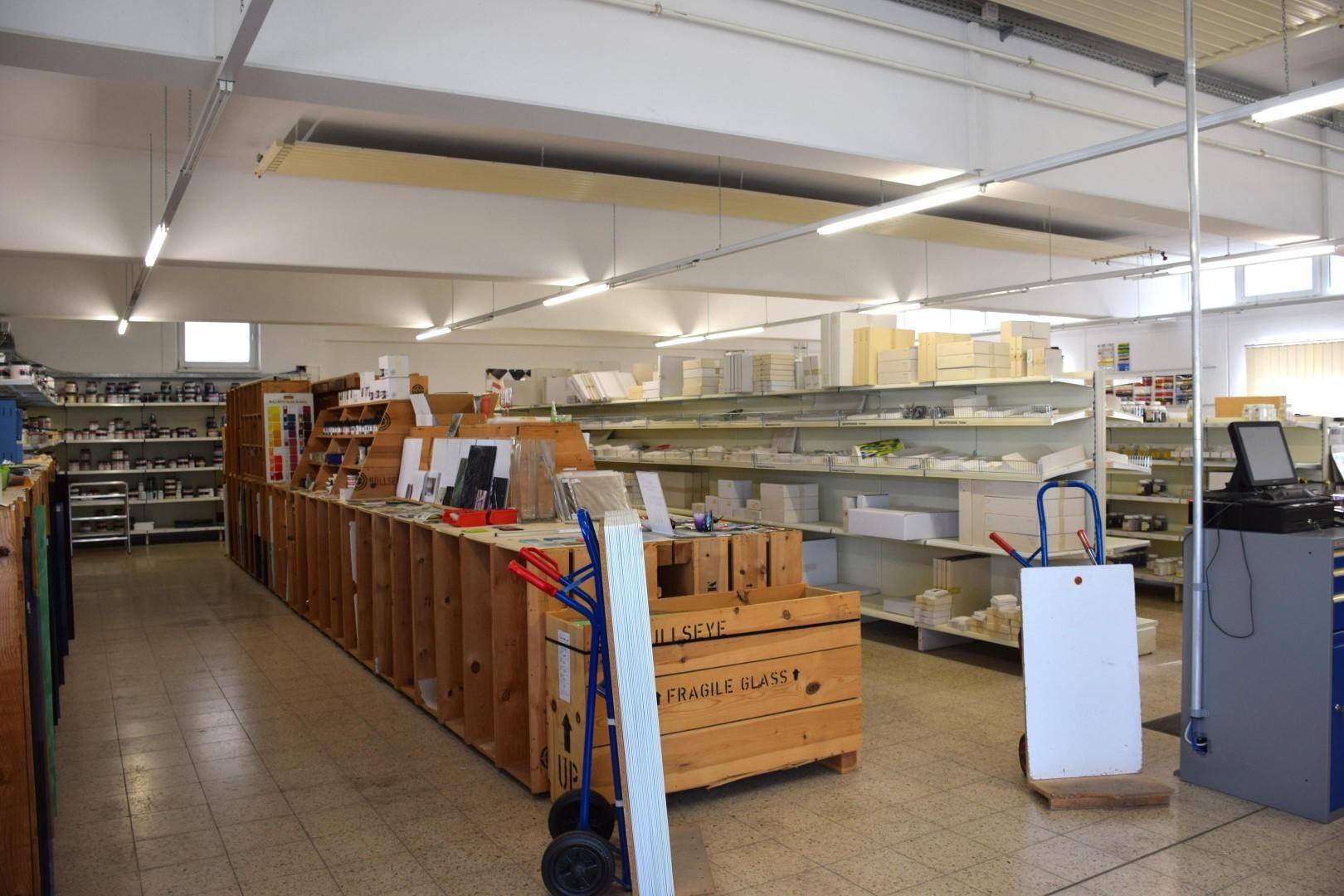Großer Lagerraum mit Regalen voller Produkte, Kisten und Arbeitsgeräten unter Leuchtstoffröhren.
