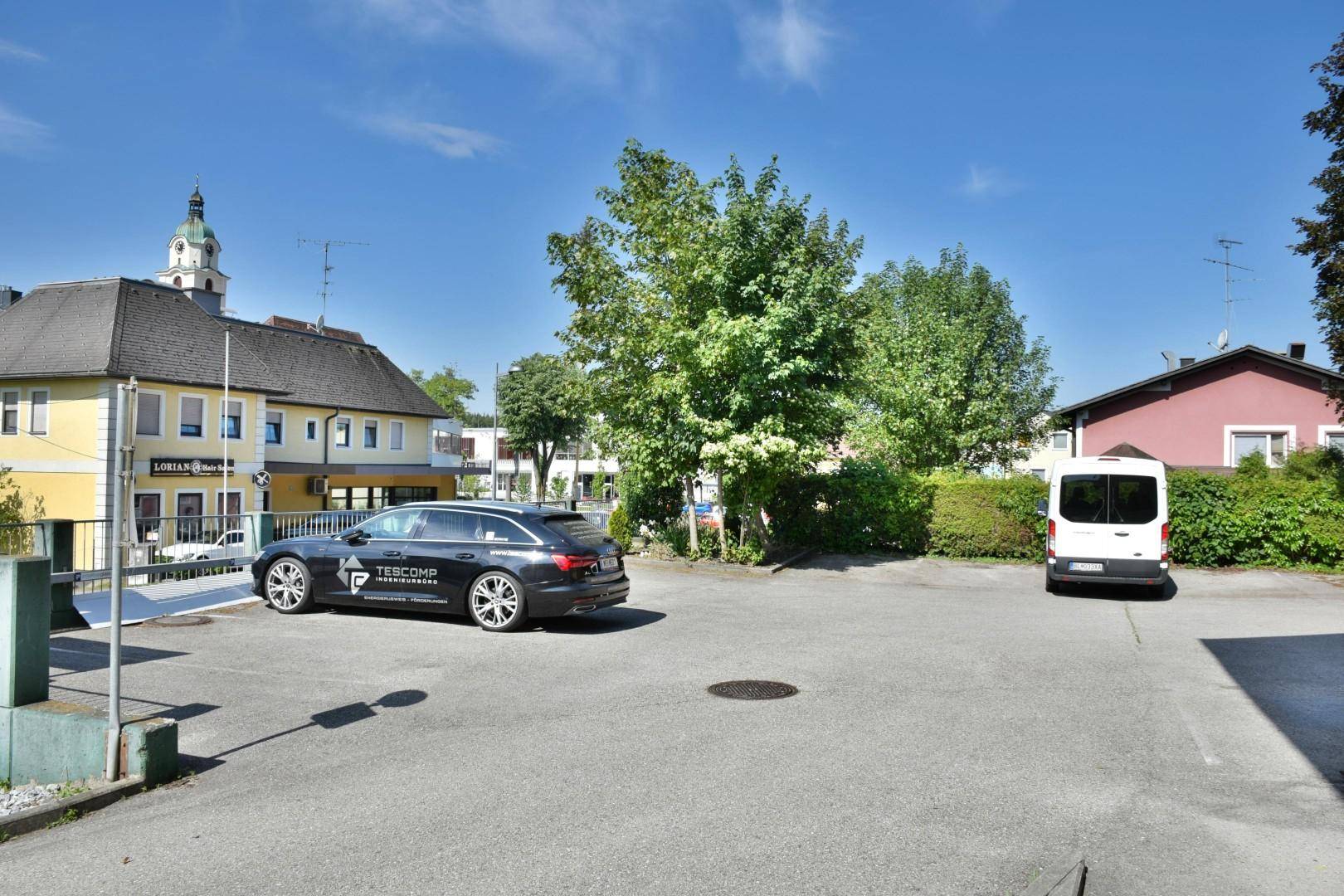Großer Außenparkplatz mit mehreren Fahrzeugen, umgeben von Bäumen und Gebäuden im Hintergrund.