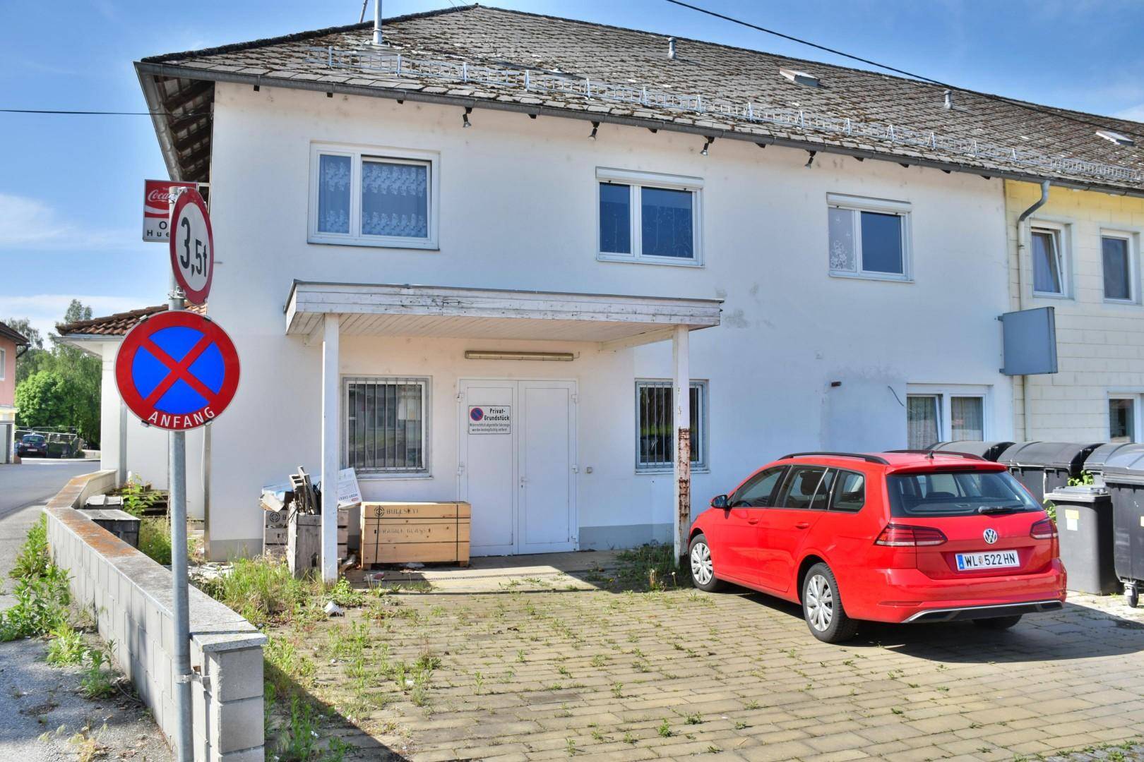 Rückseite eines Gebäudes mit gepflastertem Parkplatz, einem roten Auto und Mülltonnen.