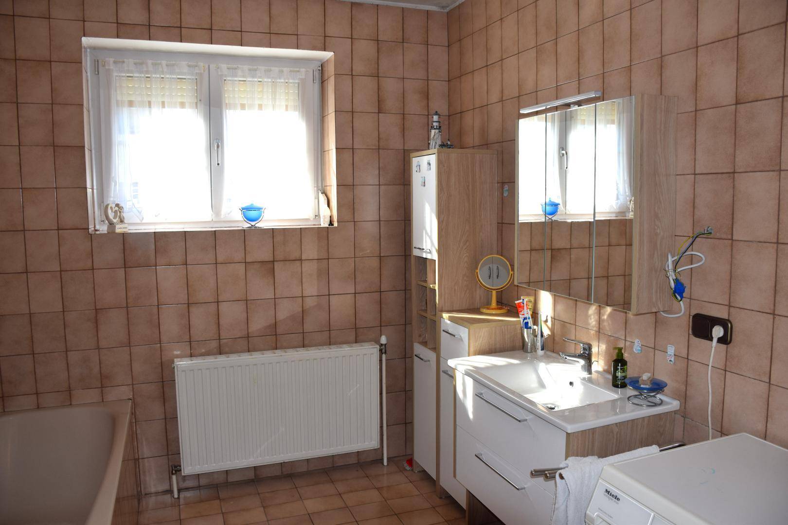 Badezimmer mit braunen Fliesen, Waschtisch, Spiegelschrank und Fenster.