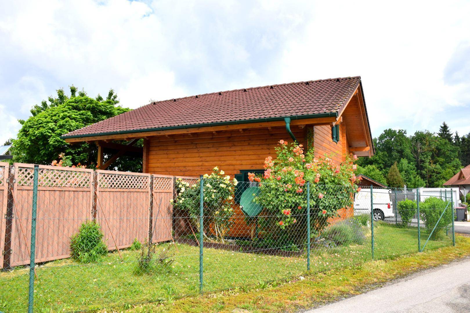 Seitlicher Blick auf das Holzhaus mit blühenden Rosenbüschen und einem umzäunten Gartenbereich.
