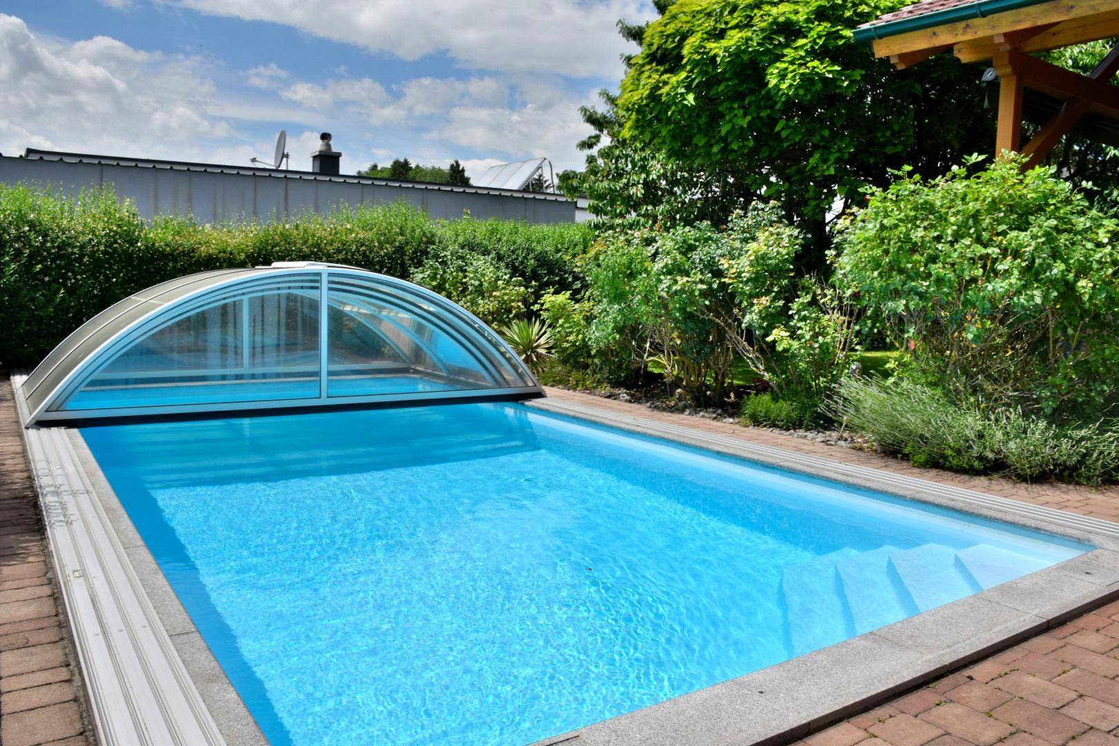 Großer Swimmingpool mit Abdeckung, umgeben von einem grünen Garten und Sträuchern.