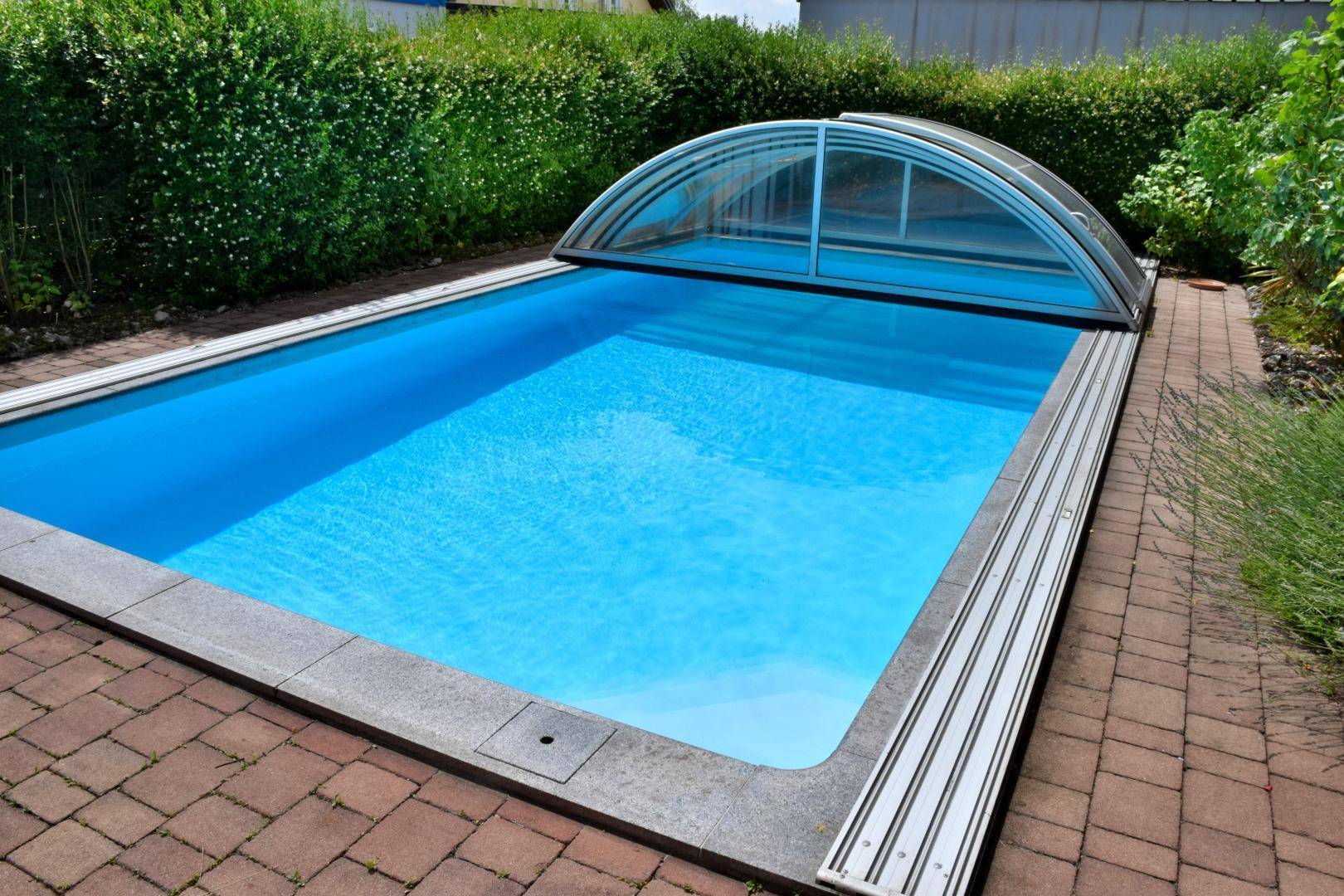 Rechteckiger Swimmingpool mit Abdeckung und gepflastertem Bereich, umgeben von Hecken.