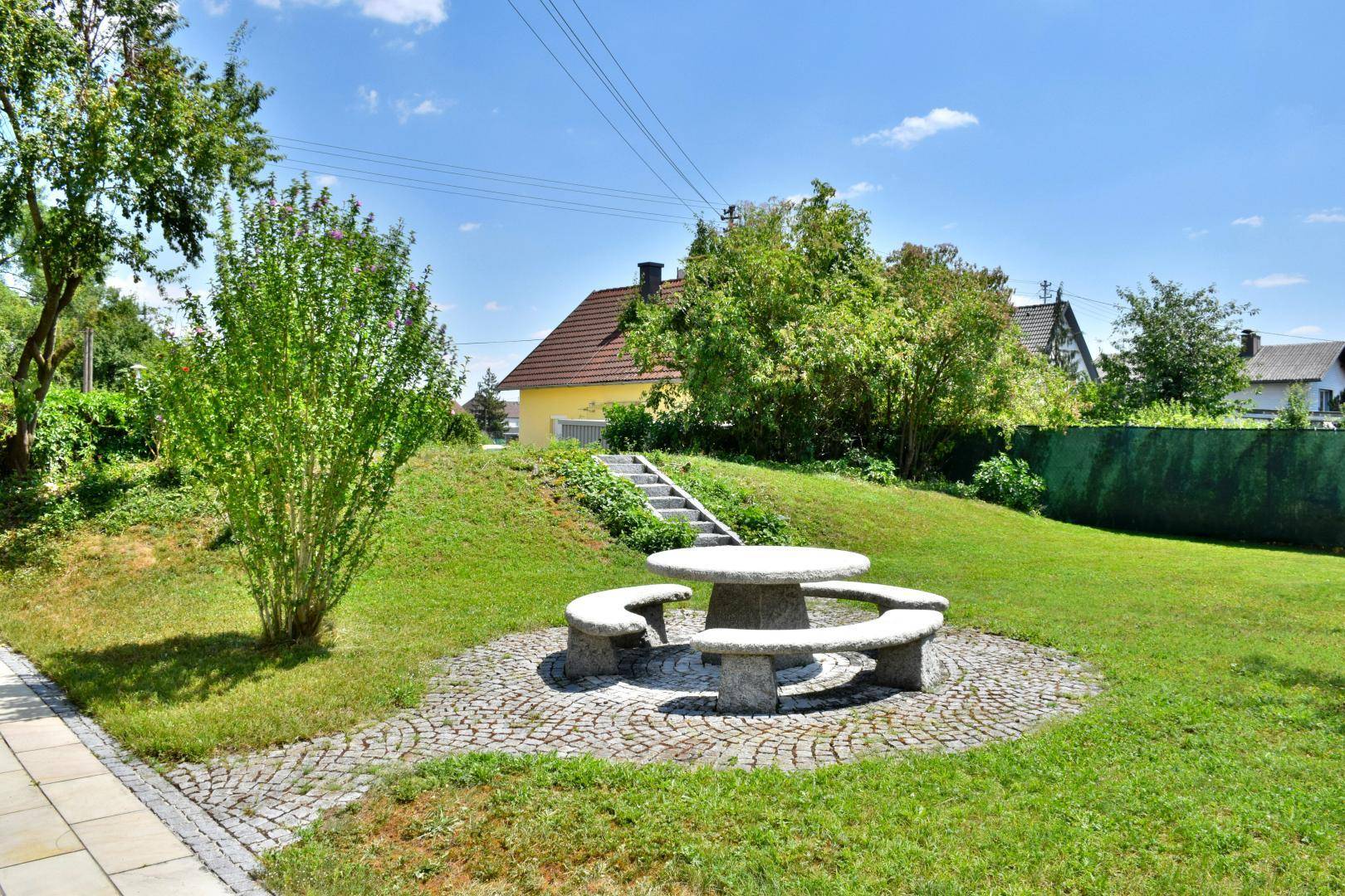Gepflasterter Bereich im Garten mit rustikalem Steintisch und Bänken, umgeben von grünen Hügeln.