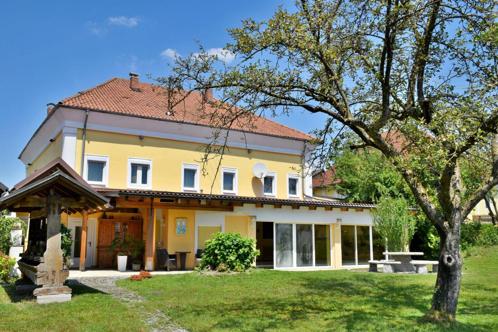 Das gelbe Wohnhaus mit rotem Dach, umgeben von einem sonnigen Garten mit altem Baum und Steintisch.