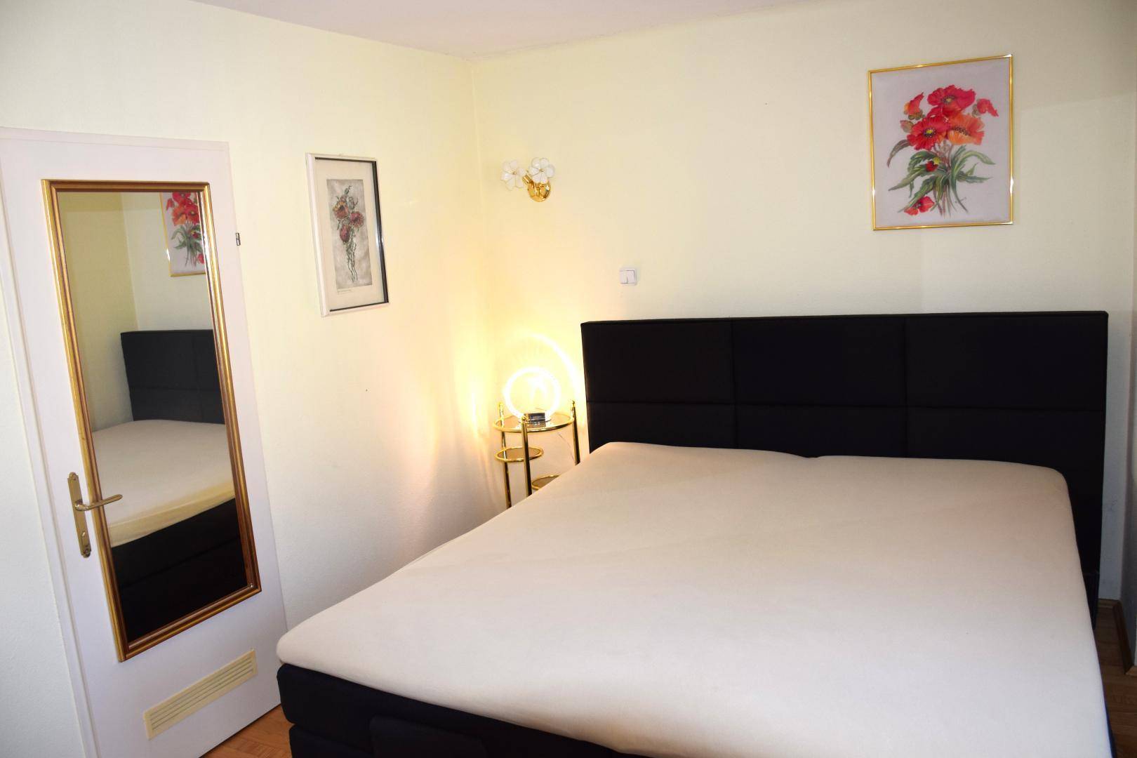 Schlichtes Schlafzimmer mit schwarzem Bett, hellem Anstrich und einem Spiegel an der Tür.