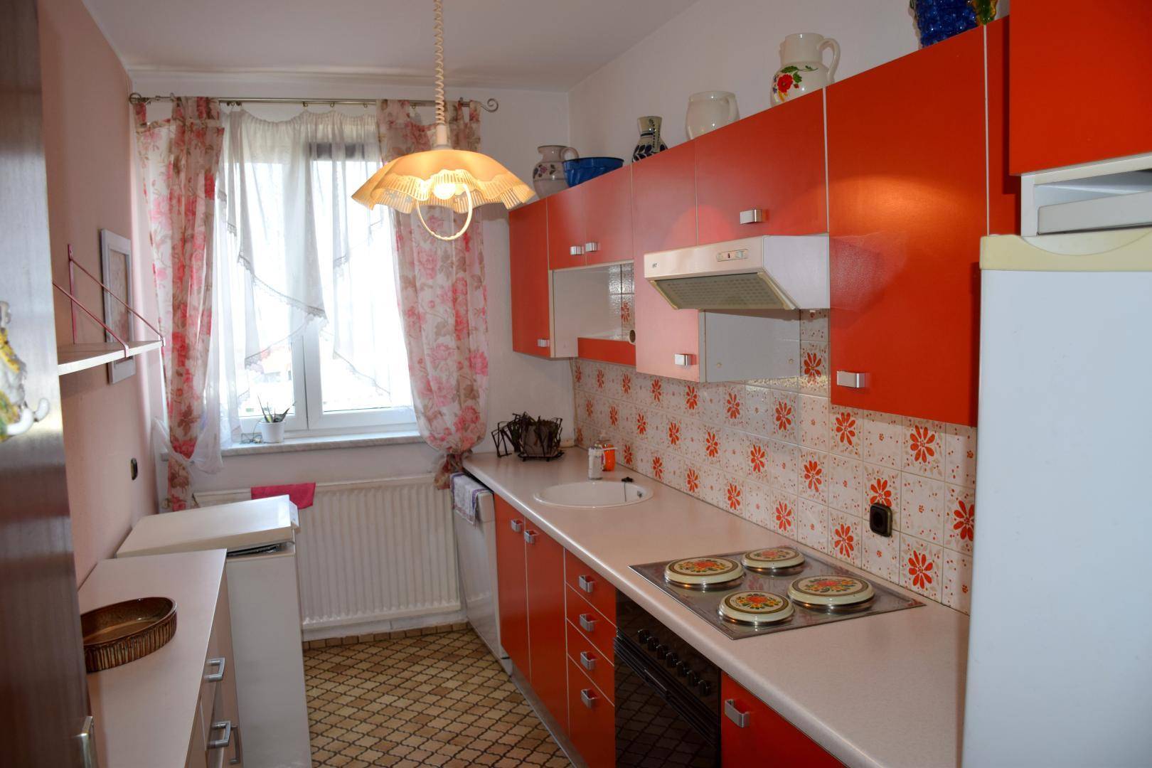 Kleine Küche mit roten Schränken, gemusterten Fliesen und Fenster mit Vorhängen.