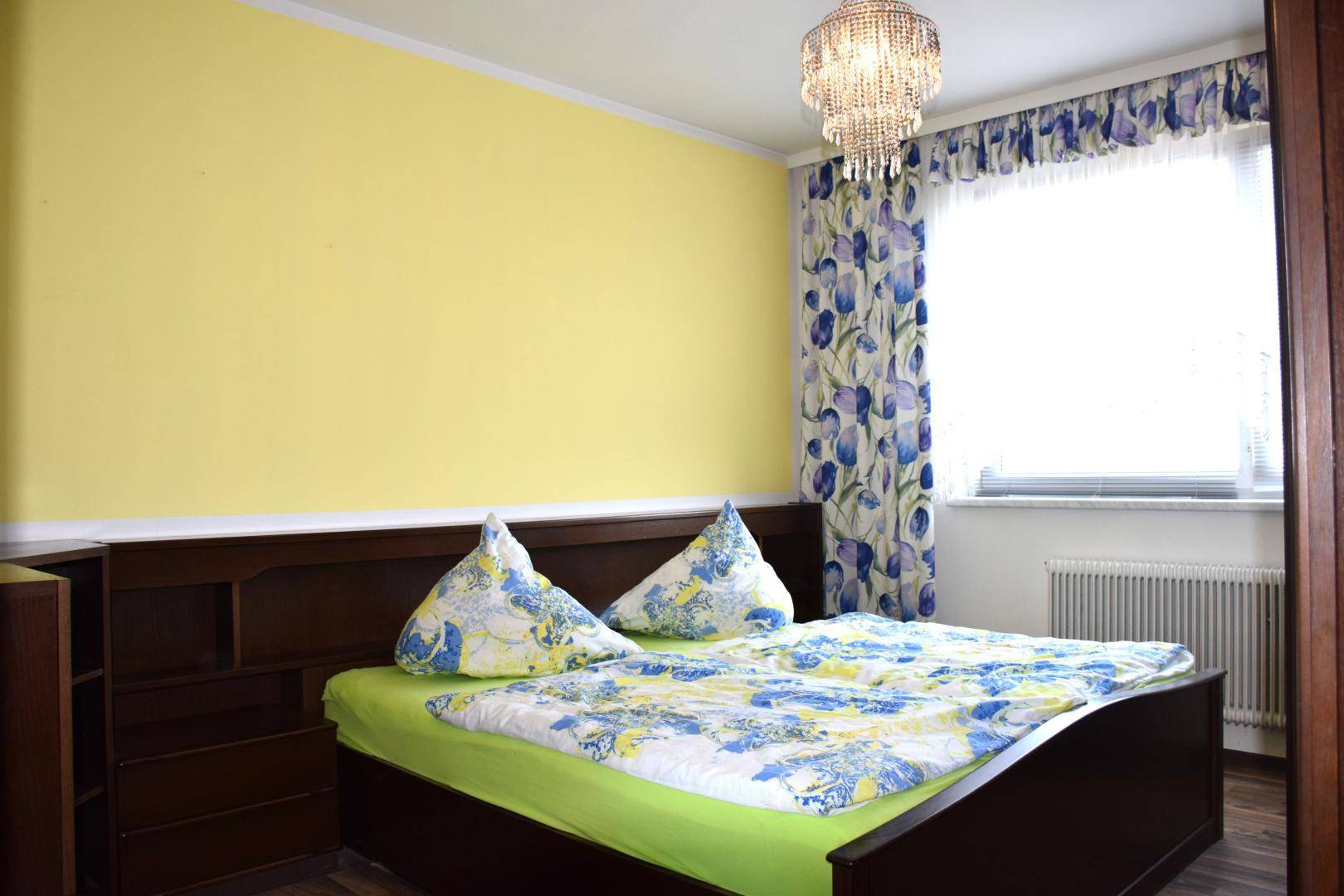 Schlafzimmer mit gelber Wand, Doppelbett, floralen Vorhängen und Kronleuchter.