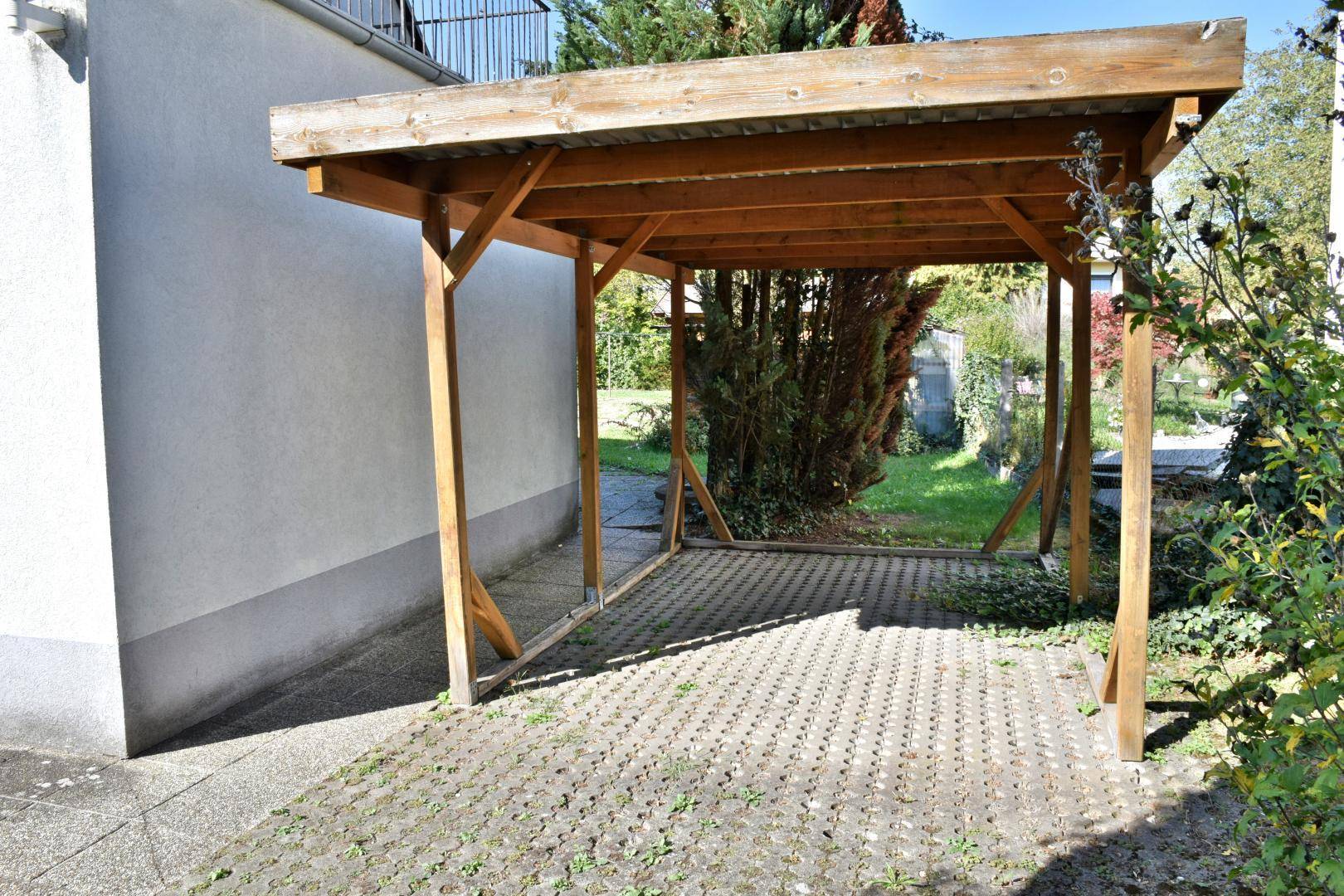 Hölzerner Carport mit gepflastertem Stellplatz und grüner Umgebung im Gartenbereich.