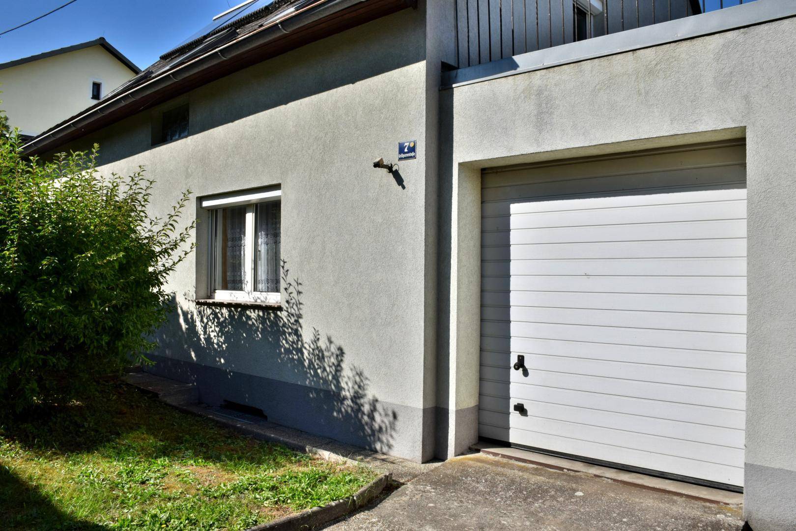 Seitenansicht des Hauses mit weißem Garagentor und einem Fenster, umgeben von Grünflächen.