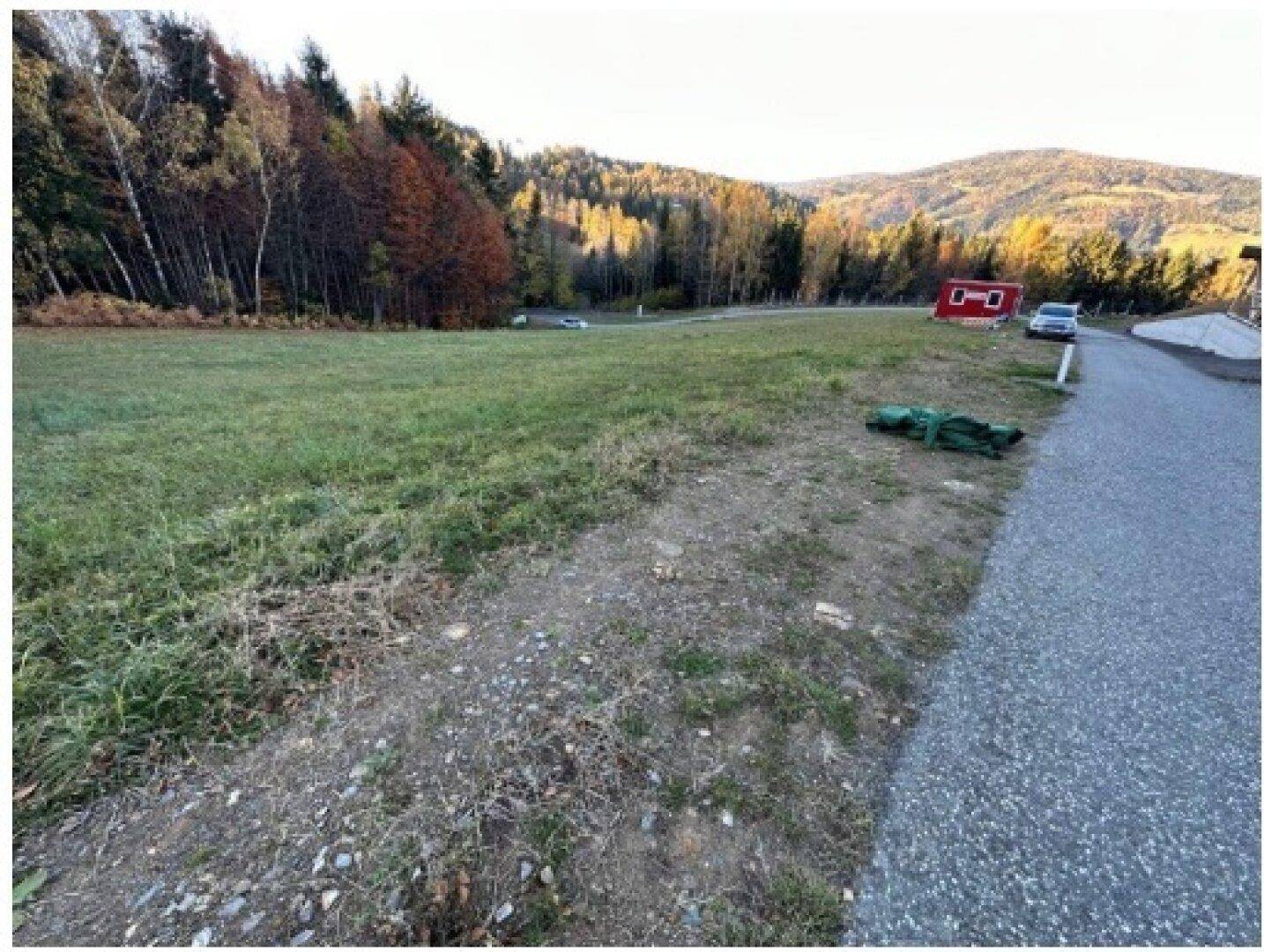 Eine weite grüne Fläche neben einer Straße, umgeben von Wäldern und Bergen im Hintergrund.