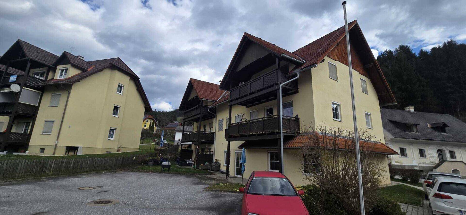 Mehrere gelbe Gebäude mit roten Dächern und Balkonen, umgeben von Bäumen und einem geparkten roten Auto.