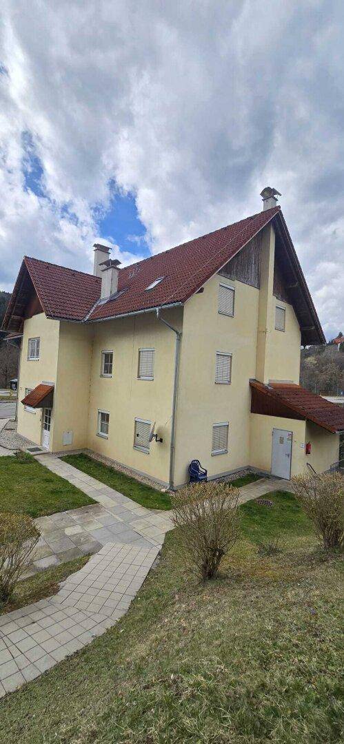 Ein gelbes Mehrfamilienhaus mit rotem Ziegeldach und grüner Umgebung unter bewölktem Himmel.