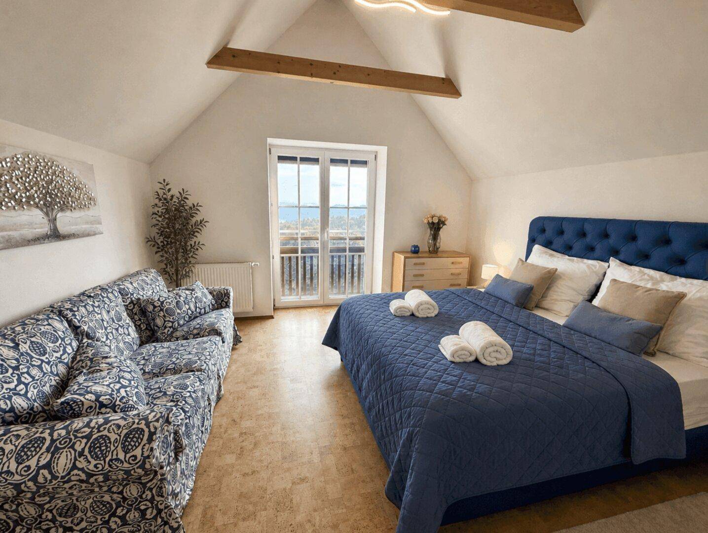 Geräumiges Schlafzimmer mit blauem Bett, Sofa und Balkonzugang, unter sichtbaren Holzbalken.