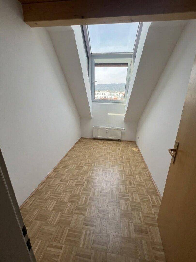 Helles Zimmer mit Parkettboden, schrägen Wänden und einem großen Dachfenster mit Ausblick.
