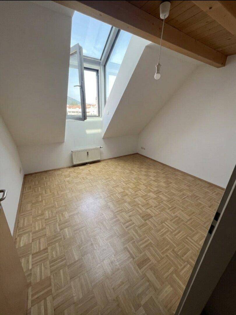 Lichtdurchflutetes Zimmer mit Parkettboden, schrägen Wänden und einem weit geöffneten Dachfenster.